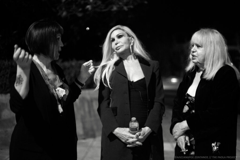 Three women stand together, engaged in conversation in a black and white photo. Wearing all black, the brunette with a chin-length bob is gesturing with her hands. The women with long blond hair is holding a bottle of water with her fingers clasped in front of her body, listening. The third woman, smoking a cigarette, is responding.