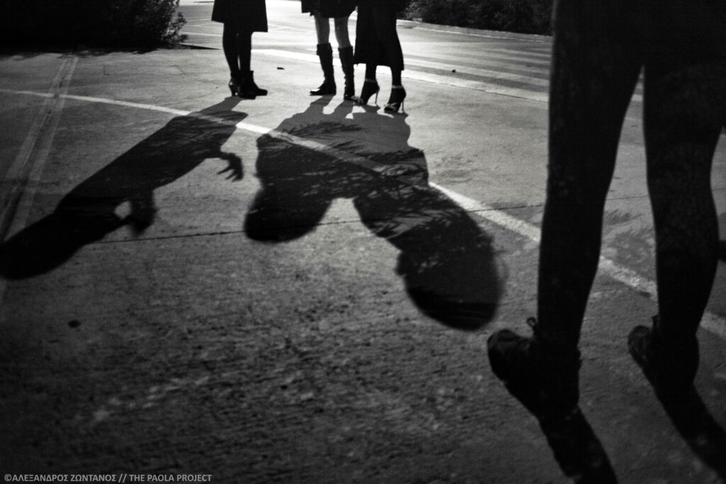 A black and white photo shows the bottom half of three people in the background, two in skirts, casting a shadow across the cement into the foreground where two legs stand in pants facing the other group.