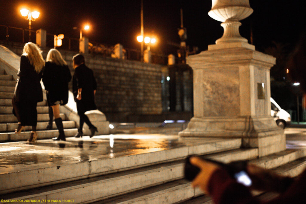 Three women in back, two with long blond hair and one brunette, climb the marble stairway. In the foreground someone is capturing their photo as they ascend the stairs toward the street lights into the dark.